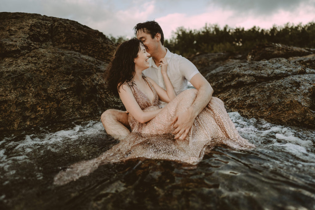 A couple is sittibng on a rock as a wave washes over them, laughing together as they get married in Vanuatu.